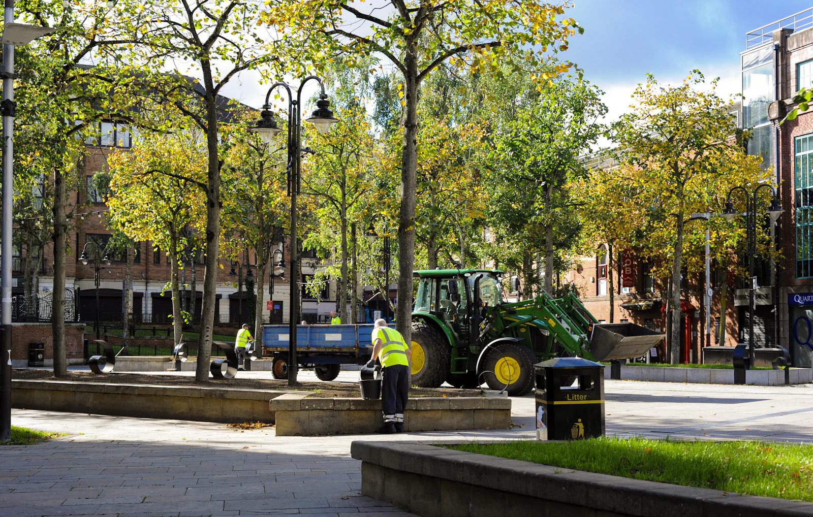 Grounds worker tending planting in a landscaped, tree-lined public plaza