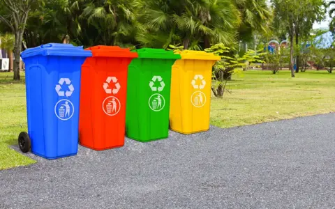 Blue, red, green and yellow recycling bins in a row beside a park lawn